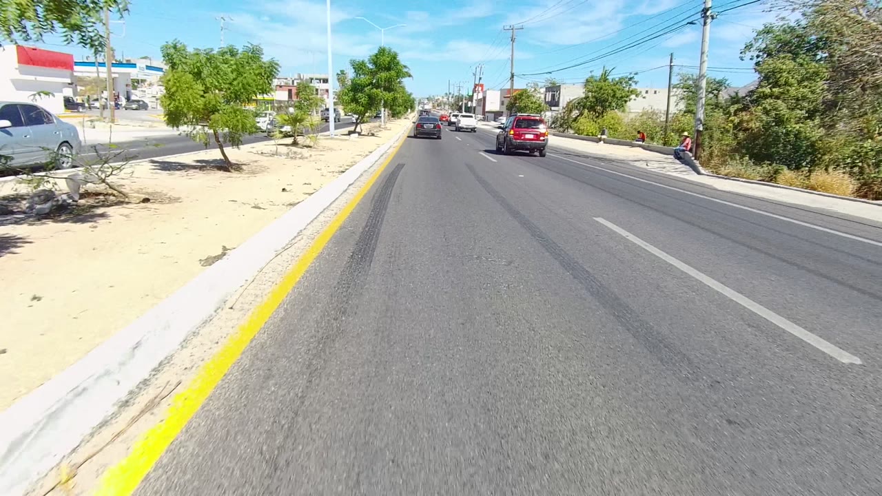 Los Cabos street view with a red SUV and other cars ahead, desert planting along the curb, overhead utility lines, and commercial buildings in strong sun, with no clearly visible people.