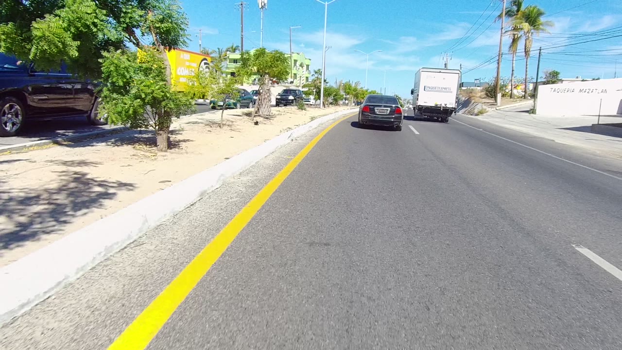 A Los Cabos street view shows a paved road, yellow curb line, parked cars under small trees, traffic ahead, sandy roadside, blue sky, and no visible people.