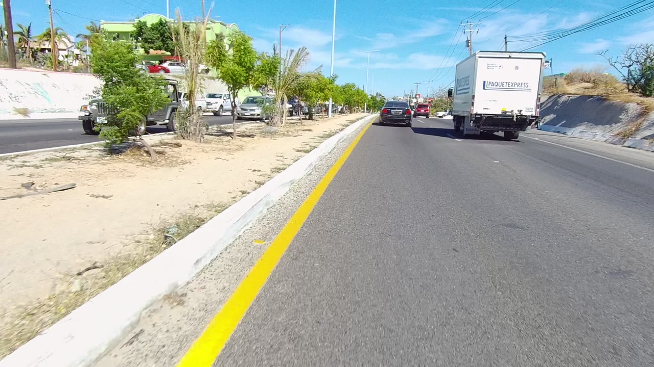 A white delivery truck and a black car travel ahead on a Los Cabos street bordered by sand, small trees, palms, low buildings, utility wires, and no visible people.