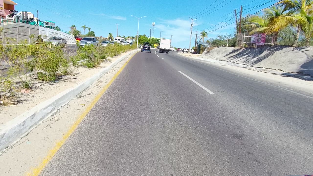 Opening frame of a Los Cabos two-lane road with cars ahead, a white box truck in the lane, palms and scrub along the roadside, and no visible people.