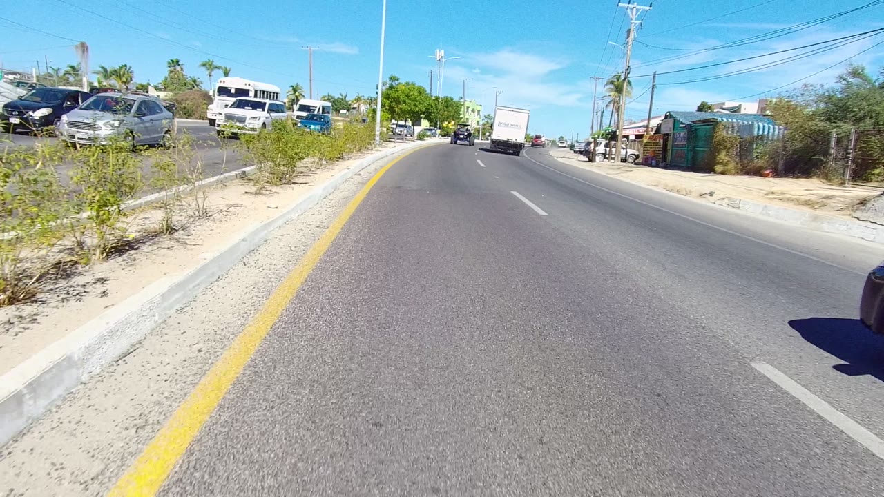 Third frame from the Los Cabos road clip, showing the lane bending slightly with a white box truck ahead, cars on both sides, low roadside buildings, palms, and no visible people.