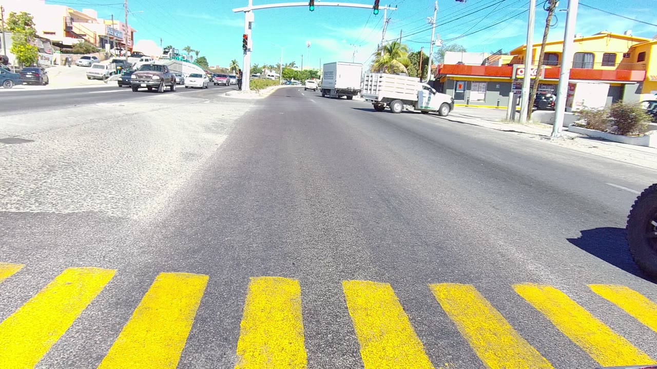 Vehicle-mounted view in Los Cabos, Baja California Sur shows yellow crosswalk bars, stopped and moving cars, traffic lights, storefronts, and no visible people at the opening moment.