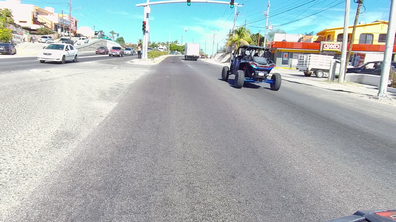 The vehicle perspective in Los Cabos shows the blue off-road vehicle farther right, with cars, a white trailer, storefronts, palm trees, traffic lights, and no visible people.