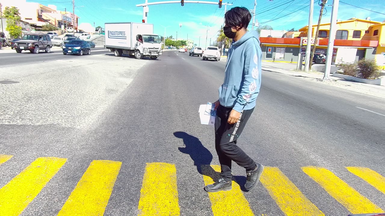 A person in a blue hoodie crosses a yellow-striped road in Los Cabos, Baja California Sur, with cars, a white box truck, storefronts, and traffic lights in bright daylight.