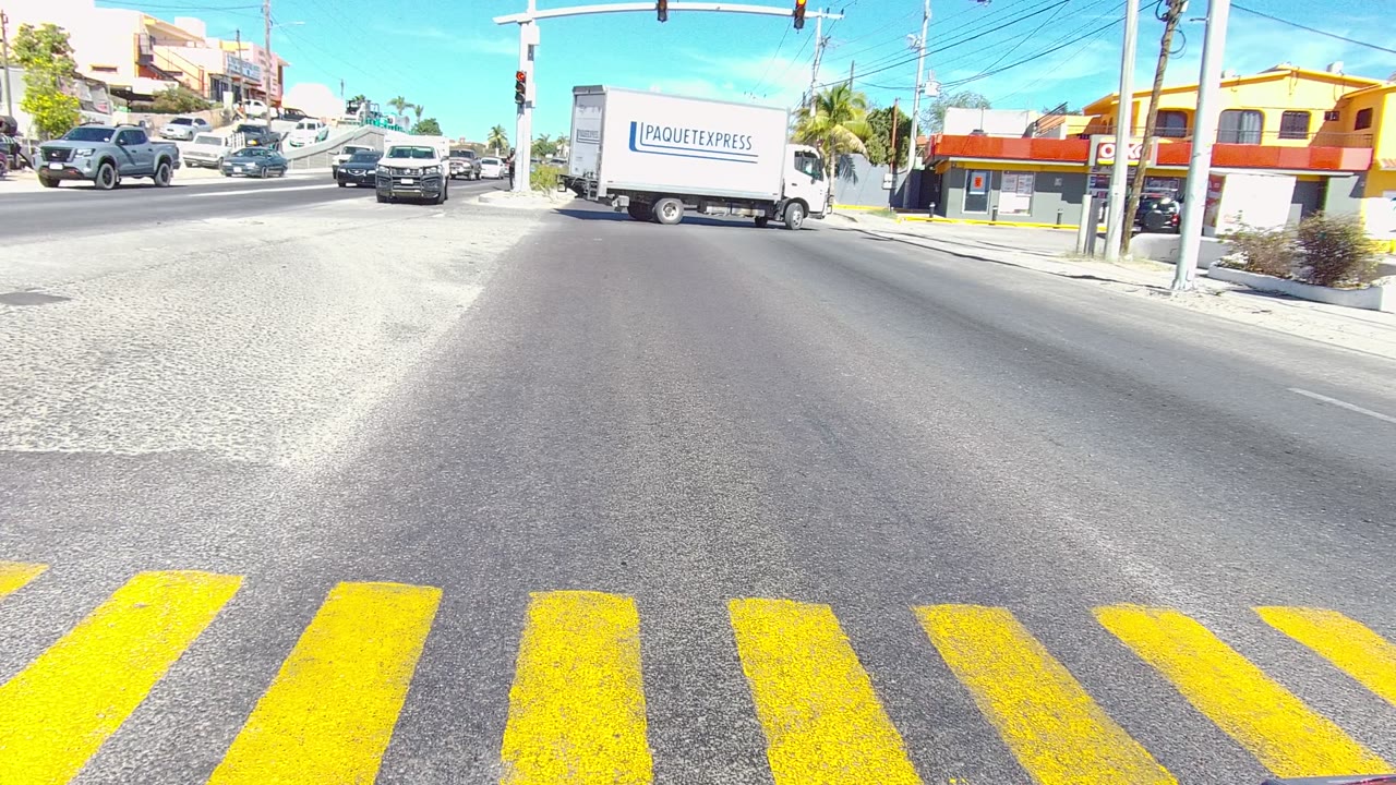No visible people are in this Los Cabos intersection frame, where a white box truck, cars, traffic signals, yellow crosswalk markings, and roadside buildings fill the view.