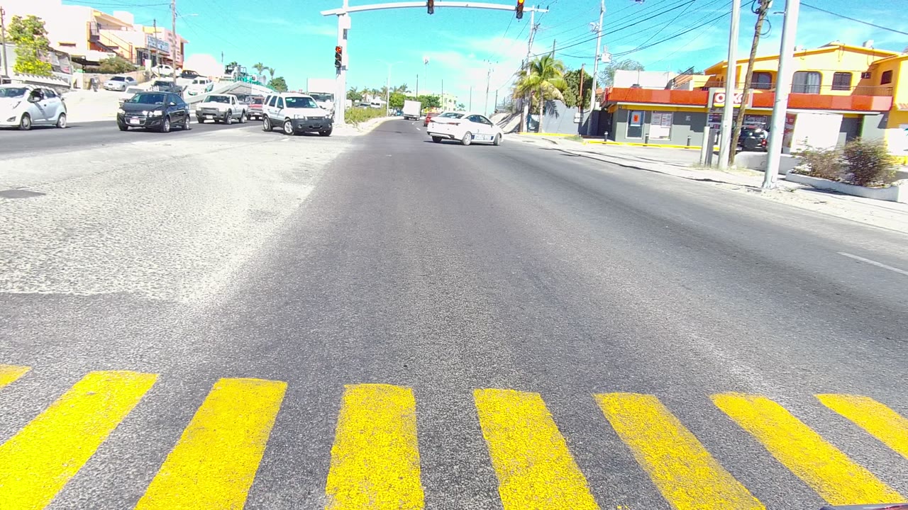 Los Cabos intersection with yellow crosswalk stripes in the foreground, several cars in the road, traffic lights overhead, low buildings at right, and no visible people.