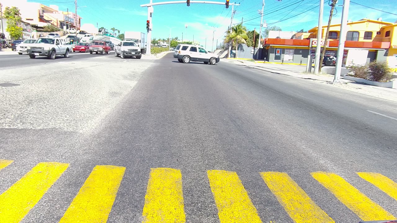 Los Cabos intersection with multiple vehicles, including a dark SUV and a white pickup, crossing the road beneath suspended traffic lights, with no visible people.