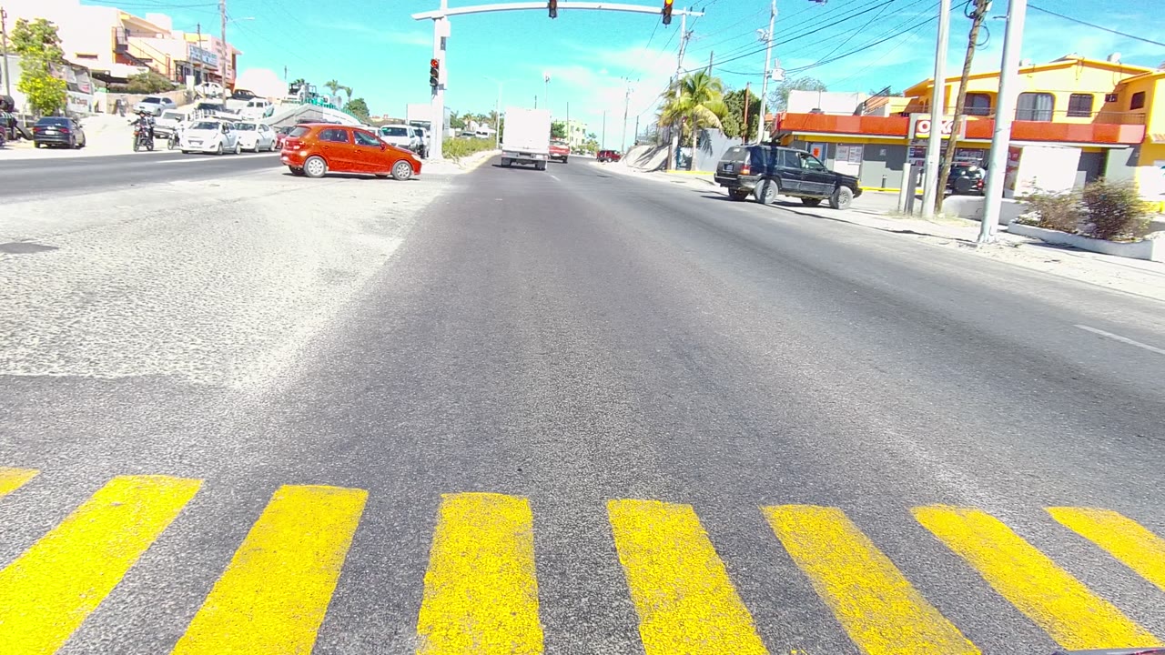 A bright Los Cabos intersection at 0:00 shows yellow crosswalk stripes close to the camera, a red car entering from the left, other vehicles on the road, storefronts, traffic lights, utility wires, and no visible people.