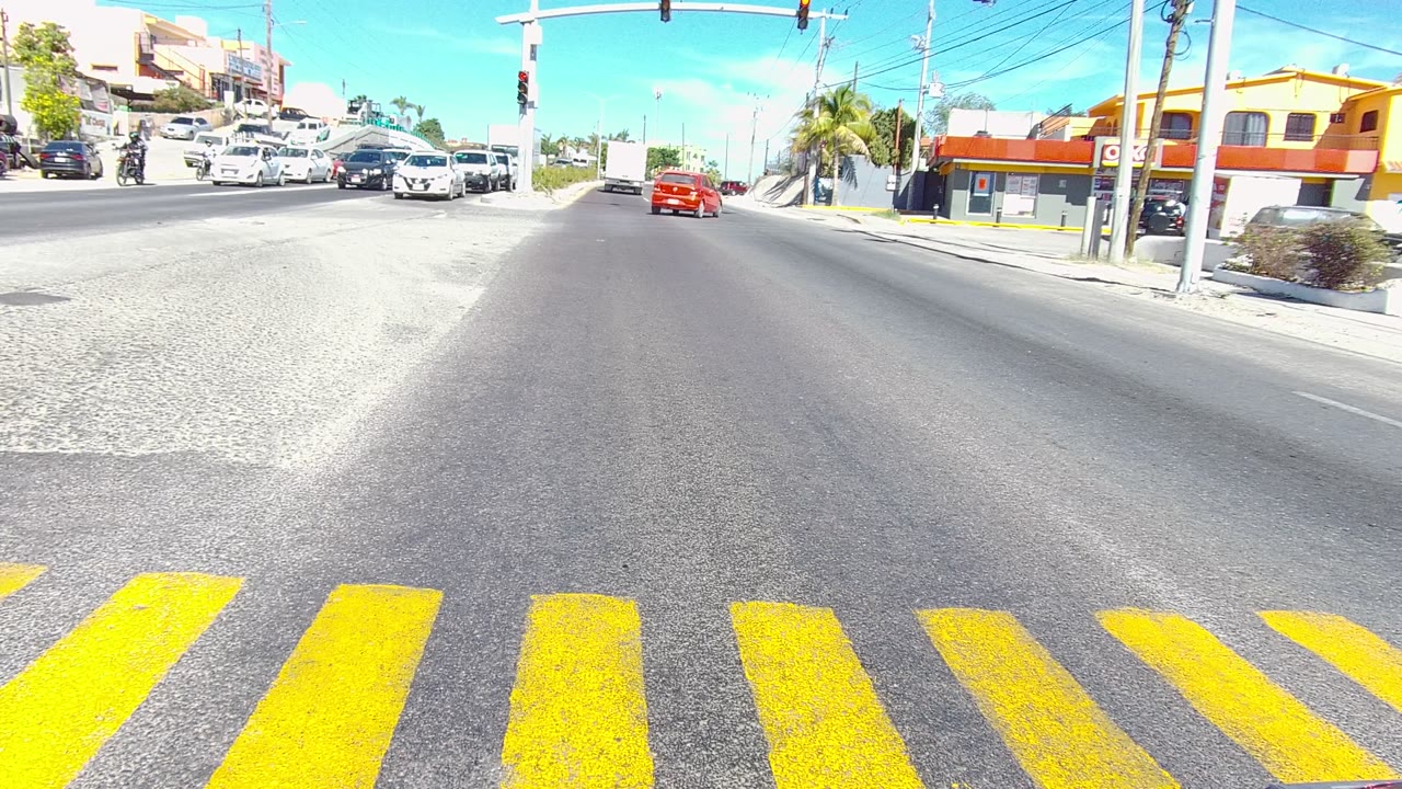 The 0:02 frame shows the red car moving away near the center of the Los Cabos roadway, yellow crosswalk lines nearest the lens, several cars at the side lanes, traffic lights overhead, and no visible people.