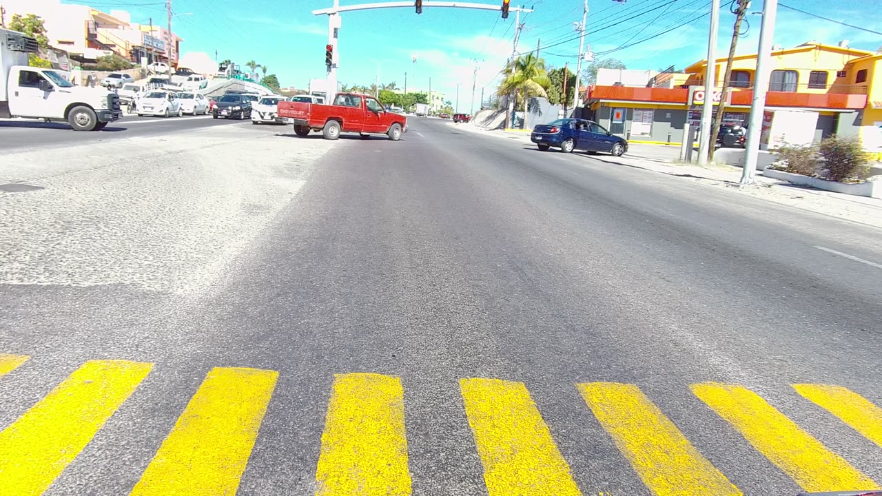 Vehicles pass through a Los Cabos street intersection beyond yellow crosswalk stripes, with buildings, power lines, overhead signals, and no visible people.