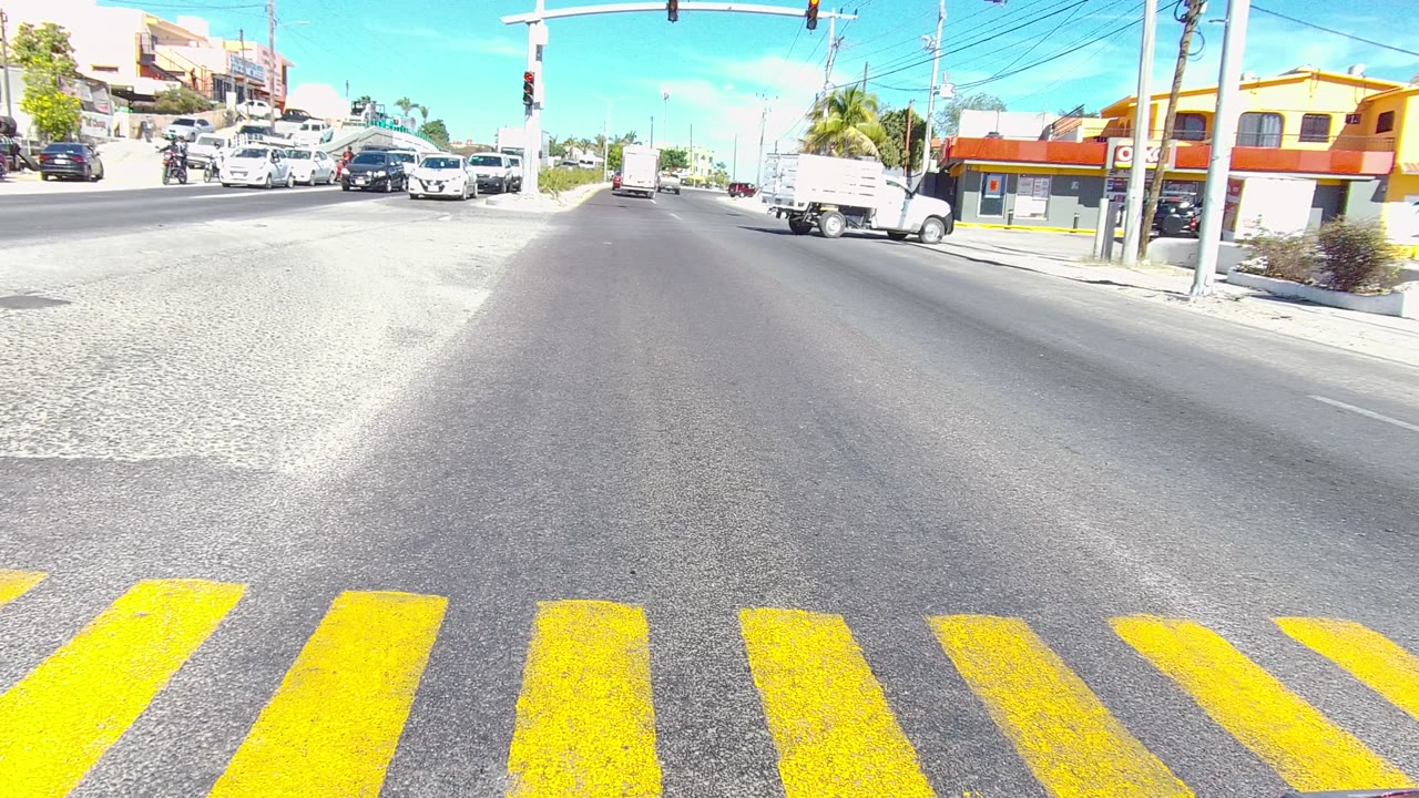 Los Cabos, Baja California Sur intersection with bright yellow crosswalk stripes in the foreground, several cars in the distance, traffic lights above, storefronts at right, and no visible people.