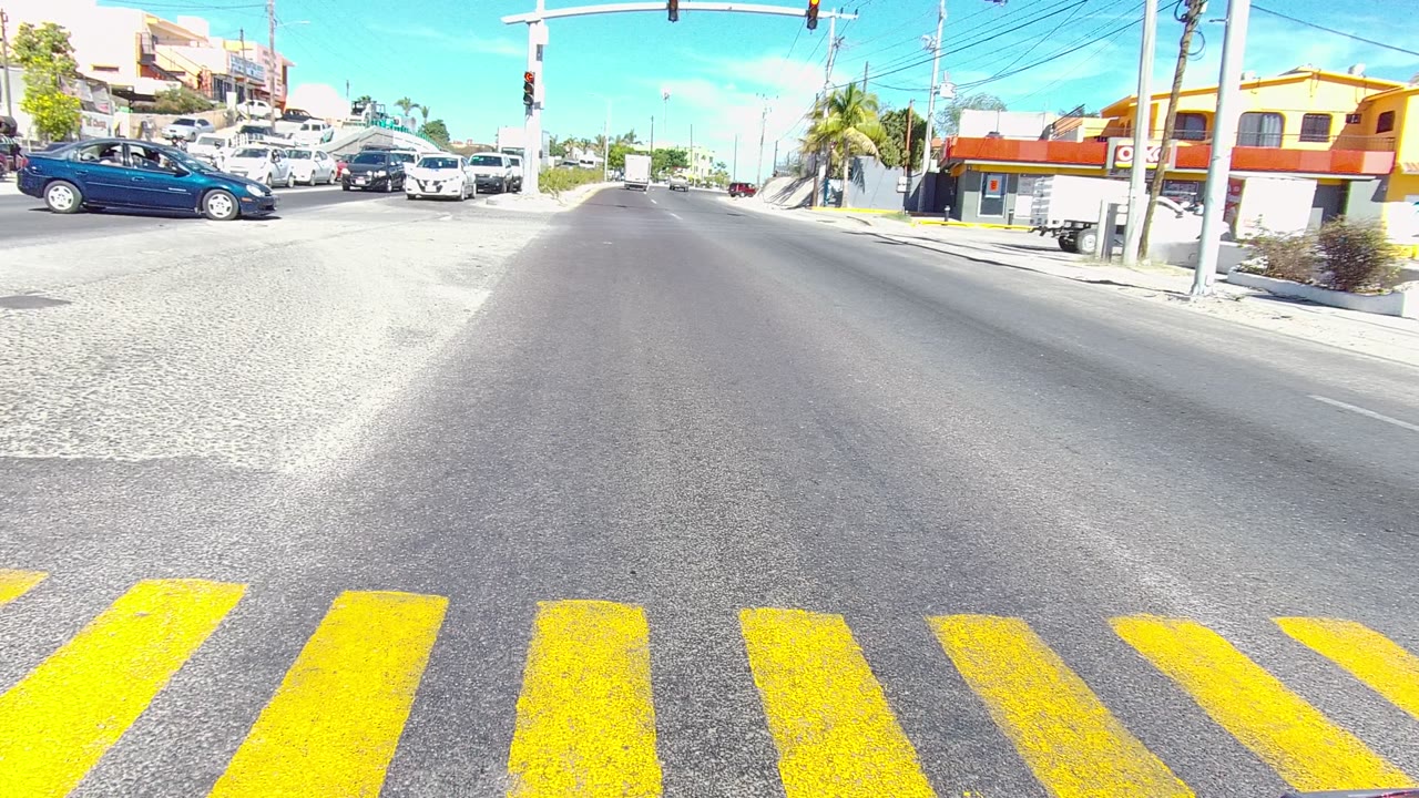 A blue car crosses the center of the Los Cabos road scene as a red pickup enters from the right, with yellow crosswalk stripes, storefronts, traffic lights, and no visible people.