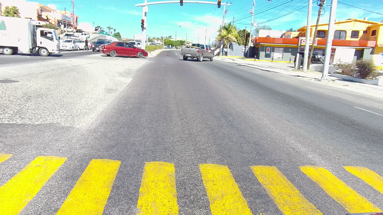 Los Cabos street intersection with yellow crosswalk stripes in the foreground, cars and trucks in traffic, traffic lights overhead, and no visible people.