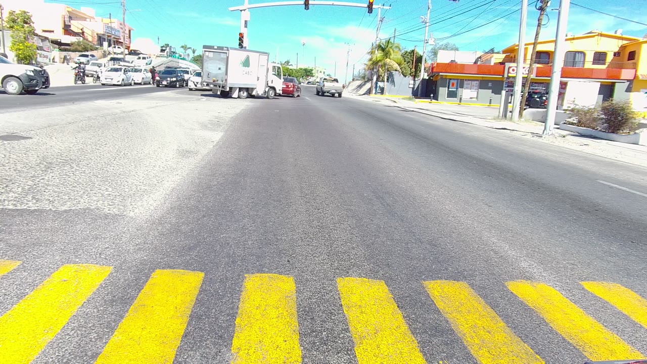 Los Cabos road junction with yellow crosswalk stripes at the bottom of frame, clustered cars and trucks across the intersection, traffic signals, and no visible people.