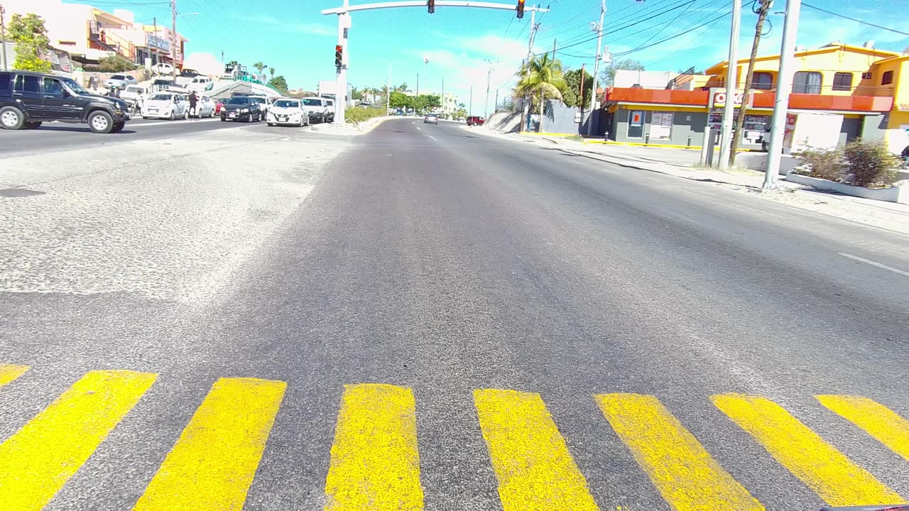 A yellow crosswalk leads into a bright Los Cabos, Baja California Sur intersection with cars waiting and driving across, low buildings in the background, and no visible people.