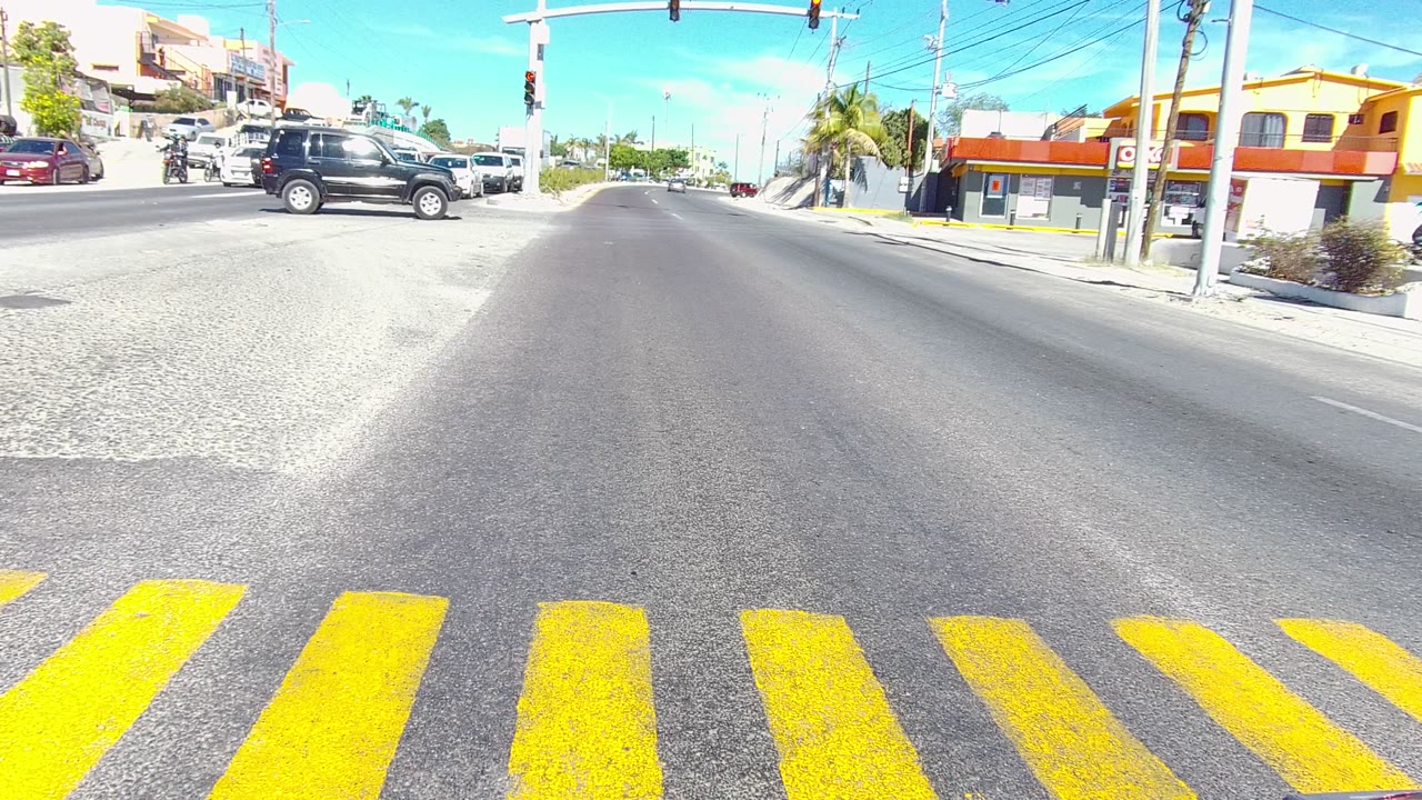 A black SUV moves across the Los Cabos intersection beyond the yellow crosswalk, with traffic lights overhead, roadside buildings to the right, and no visible people.
