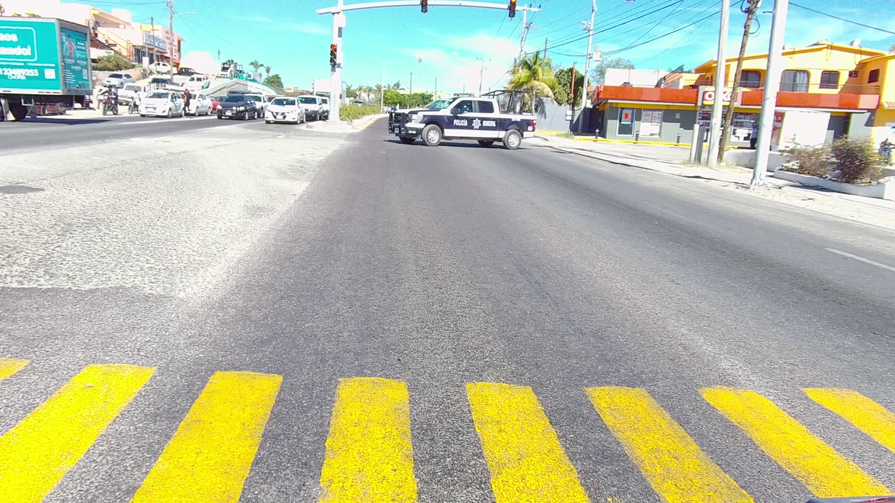 A police pickup moves through a Los Cabos, Baja California Sur intersection with yellow crosswalk stripes in the foreground, traffic lights overhead, cars nearby, storefronts behind, and no visible people.