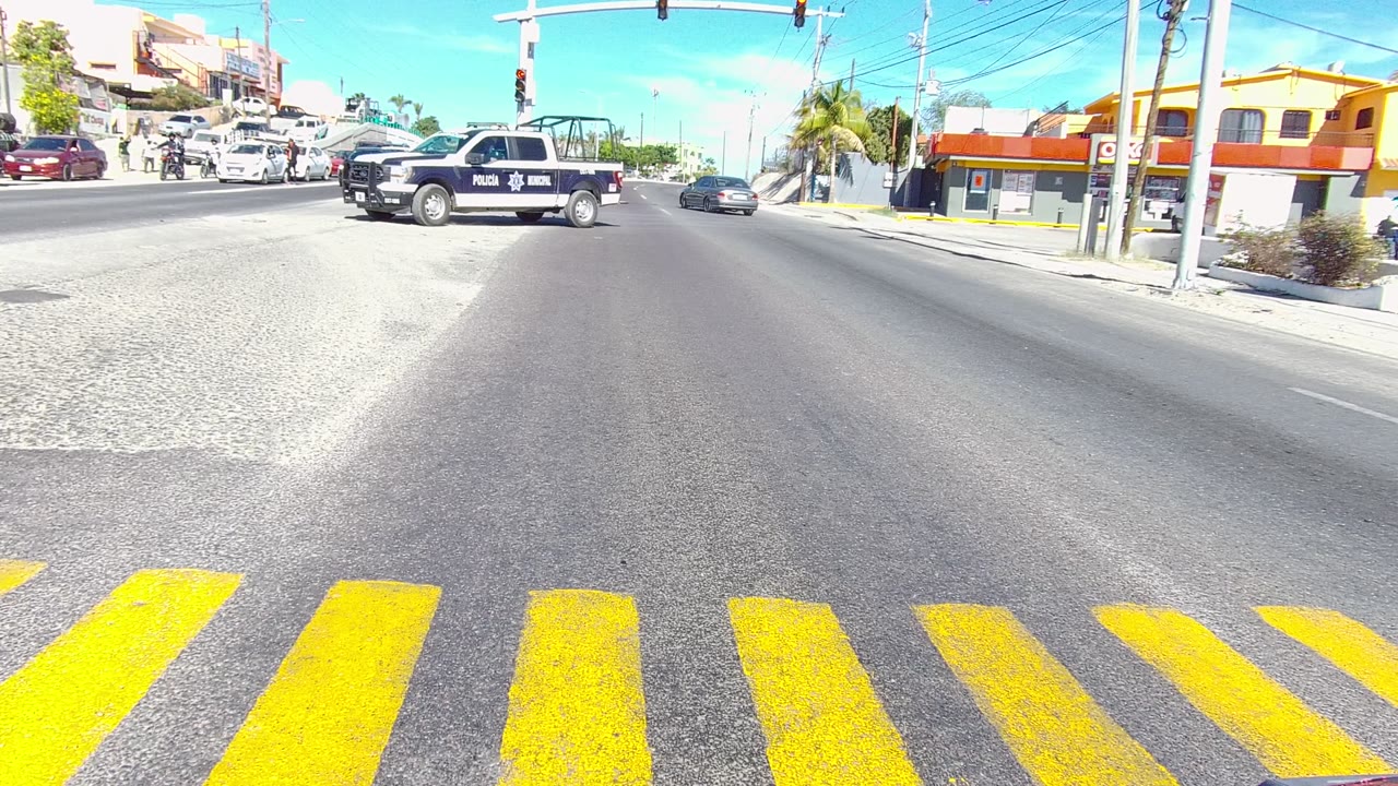 The police pickup is centered in the Los Cabos street intersection, with broad yellow crosswalk markings at the bottom of frame, cars along the cross street, traffic signals, buildings, and no visible people.