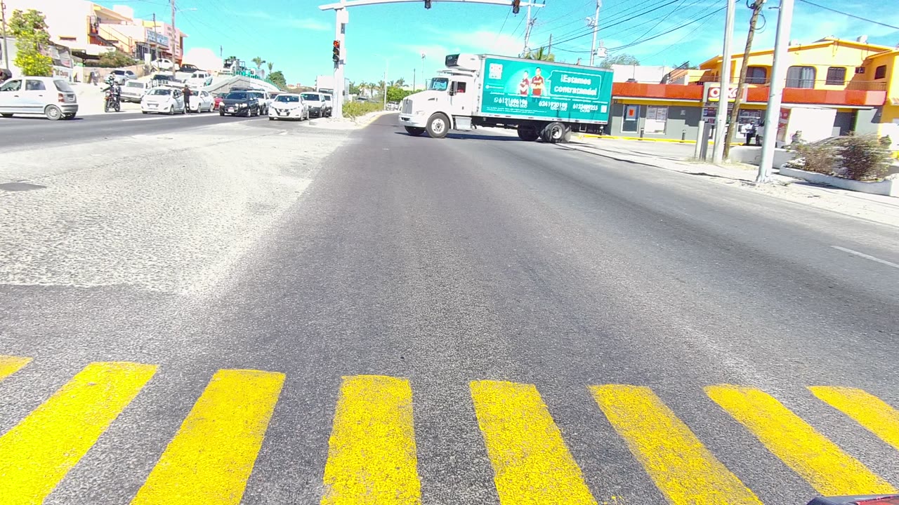 At 0:00 in Los Cabos, Baja California Sur, yellow crosswalk stripes fill the foreground as cars and a white truck with a green box move through a sunlit intersection, with no visible people.