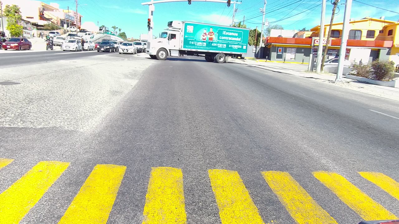 A green-sided truck crosses the intersection while cars wait and pass nearby.