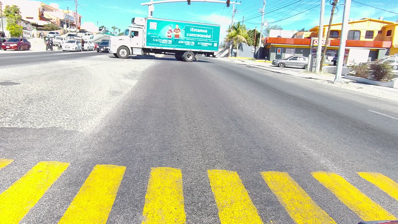 At 0:02 in Los Cabos, Baja California Sur, the white cab and green cargo box pass through the sunlit intersection, with cars behind it, yellow crosswalk markings in front, roadside buildings, and no visible people.
