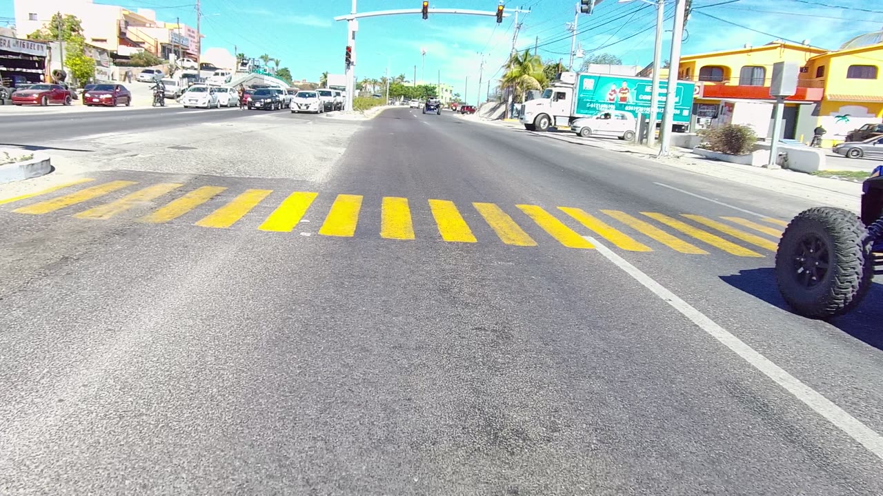 Low road-level view of a yellow crosswalk at a Los Cabos intersection with cars and trucks ahead under clear blue sky, no visible people.