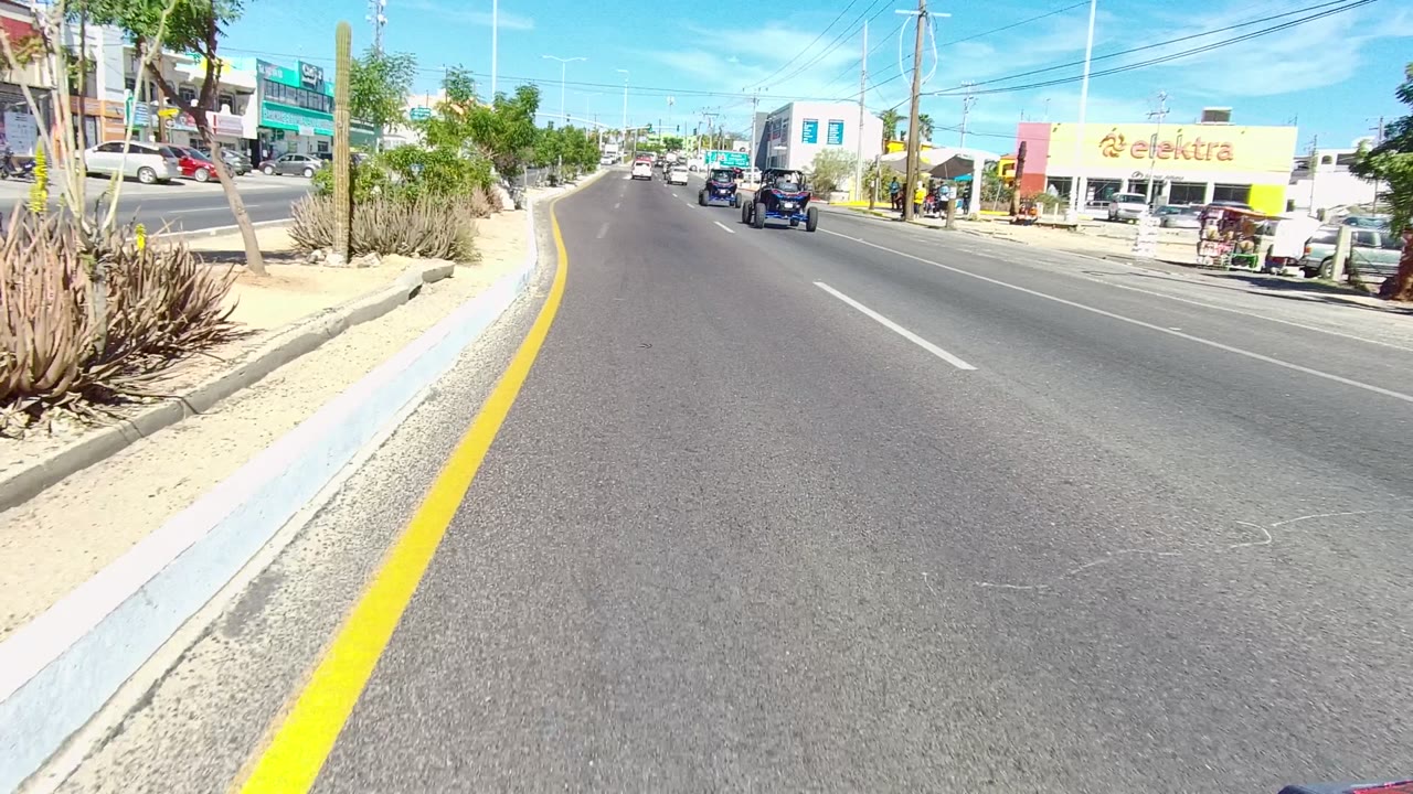 Wide road view in Los Cabos with sparse traffic ahead, desert plants along the median edge, commercial buildings, utility poles, strong sun, and no visible people in this frame.