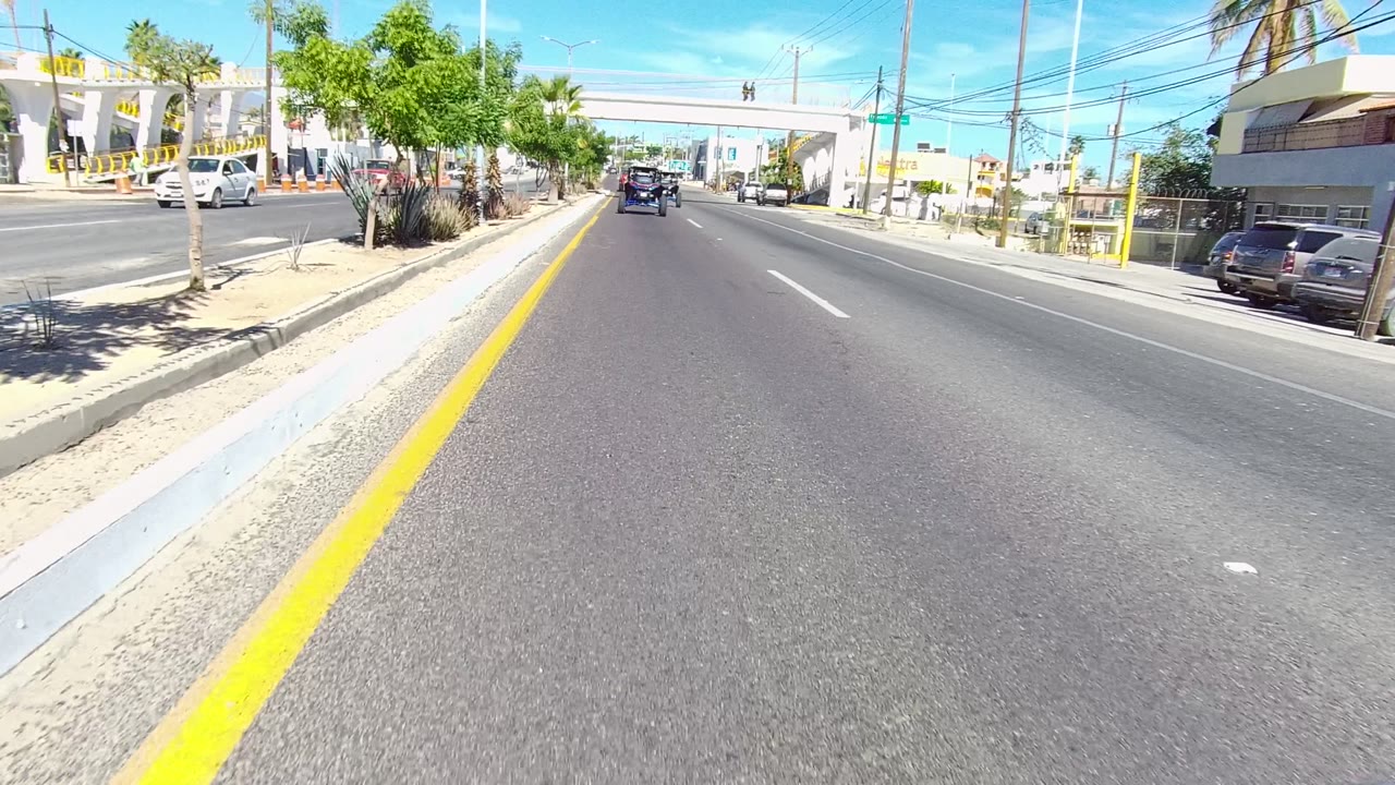 Mid-clip Los Cabos road frame with a dark vehicle ahead in the lane, storefront signage on the right, parked cars, small trees, palms, overhead wires, and no visible people at street level.