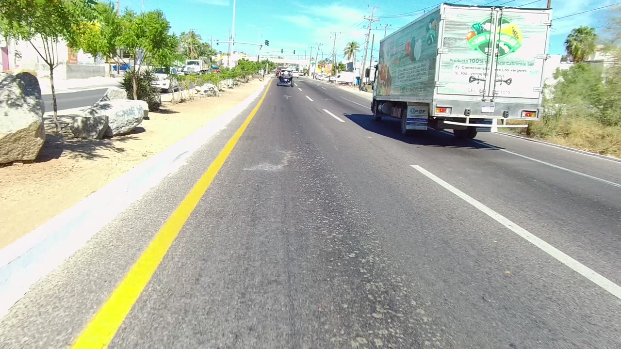 Forward road view in Los Cabos, Baja California Sur, with a large truck close on the right, yellow lane marking on the left, sidewalks, trees, buildings, and no clearly visible people.