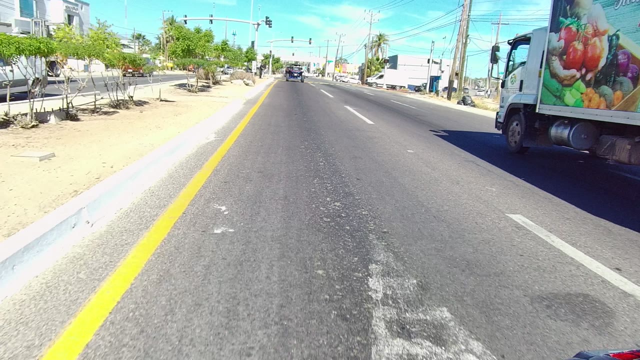 Forward-facing Los Cabos street view with the truck nearing the right edge of frame, a wide asphalt lane, sidewalk planting, commercial buildings, overhead signals, and no clearly visible people.