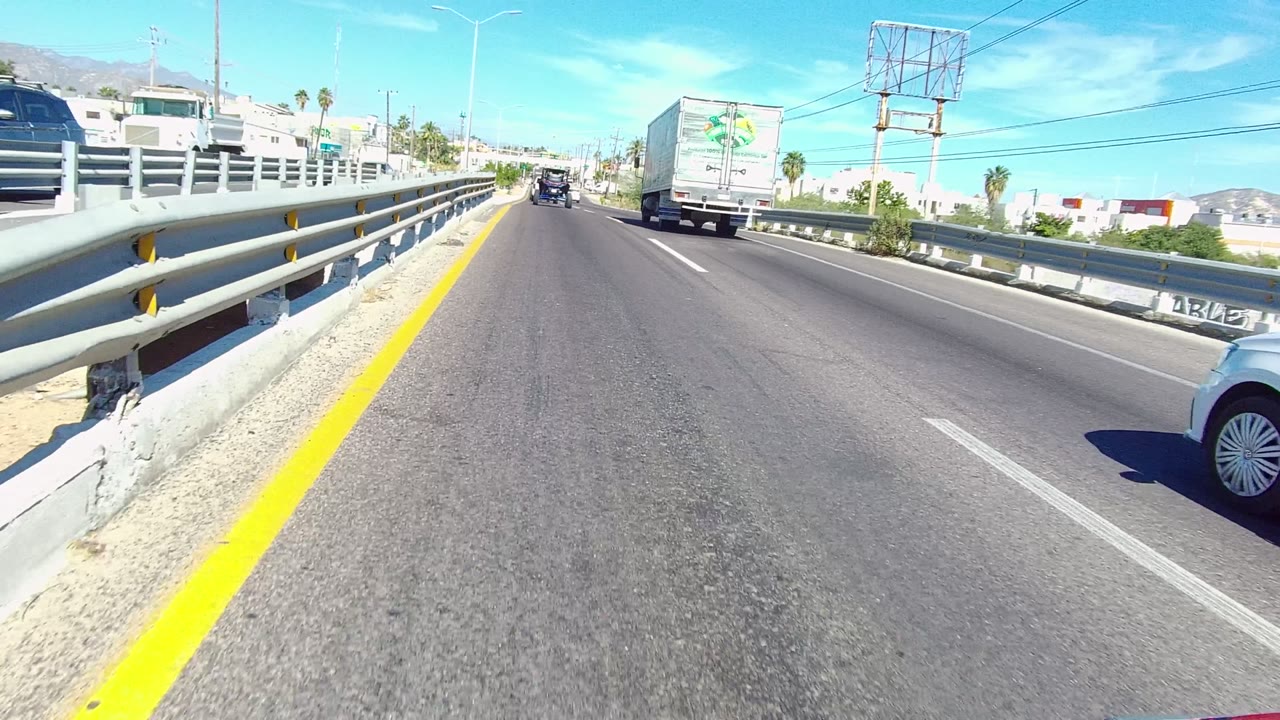 Forward-driving view on a multi-lane road in Los Cabos with metal guardrails, a white truck ahead, a dark vehicle farther forward, bright sky, and no visible people.