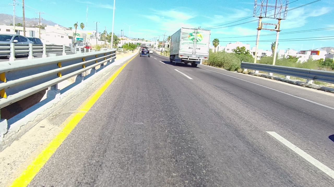 A Los Cabos road view continues past the bridge guardrail, with a white box truck in the right lane, a dark car ahead, roadside buildings, utility wires, and no visible people.
