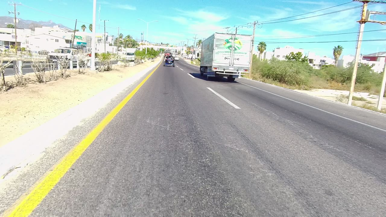 The road opens in Los Cabos with the guardrail mostly gone, showing a white truck to the right, a dark vehicle ahead, utility poles, low buildings, dry roadside ground, and no visible people.