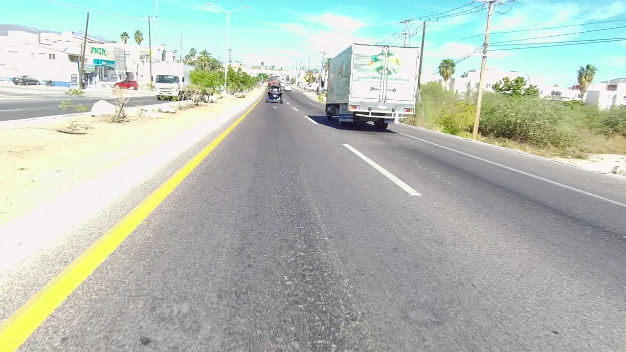 White truck ahead on a sunlit multi-lane road with buildings and vegetation.