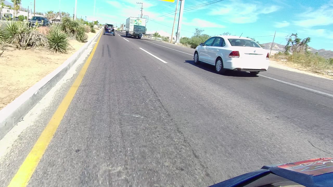 Vehicle-level road view in Los Cabos, Baja California Sur, with a white sedan to the right, a truck ahead, dry roadside plants, bright blue sky, and no visible people.