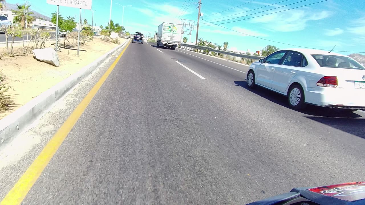 A bright Los Cabos driving frame shows the white car partly cropped at right, the box truck farther ahead, dry sandy shoulder, roadside signs, and no visible people.