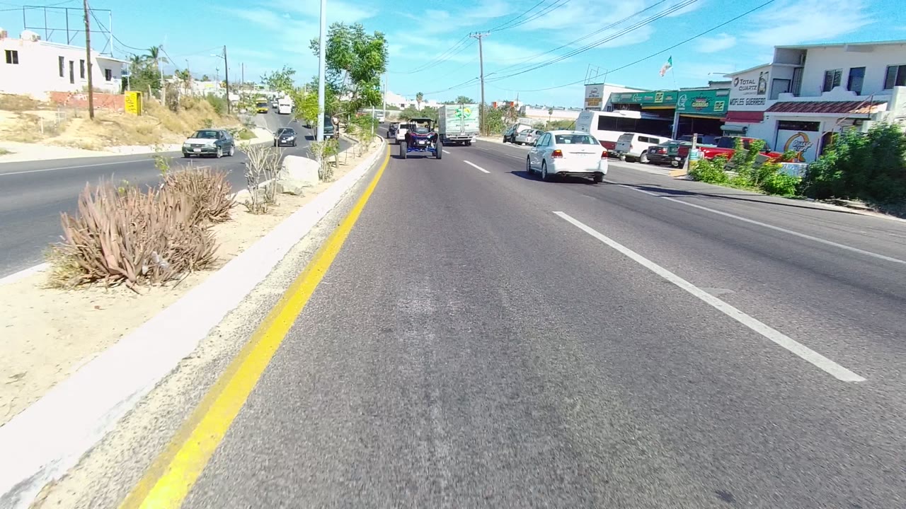 Los Cabos road view at the start of the clip, showing asphalt lanes, a sandy planted median, roadside buildings, cars and trucks ahead, and no visible people.