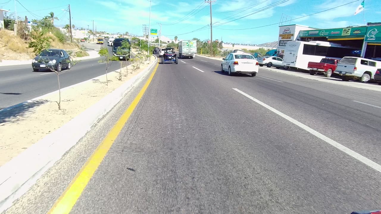 Road traffic passes businesses and a large dark bus under bright sky.