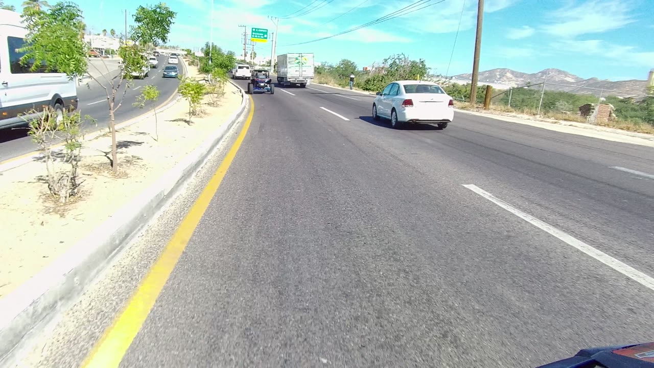 Divided road with vehicles, sandy median, roadside trees, and distant hills.
