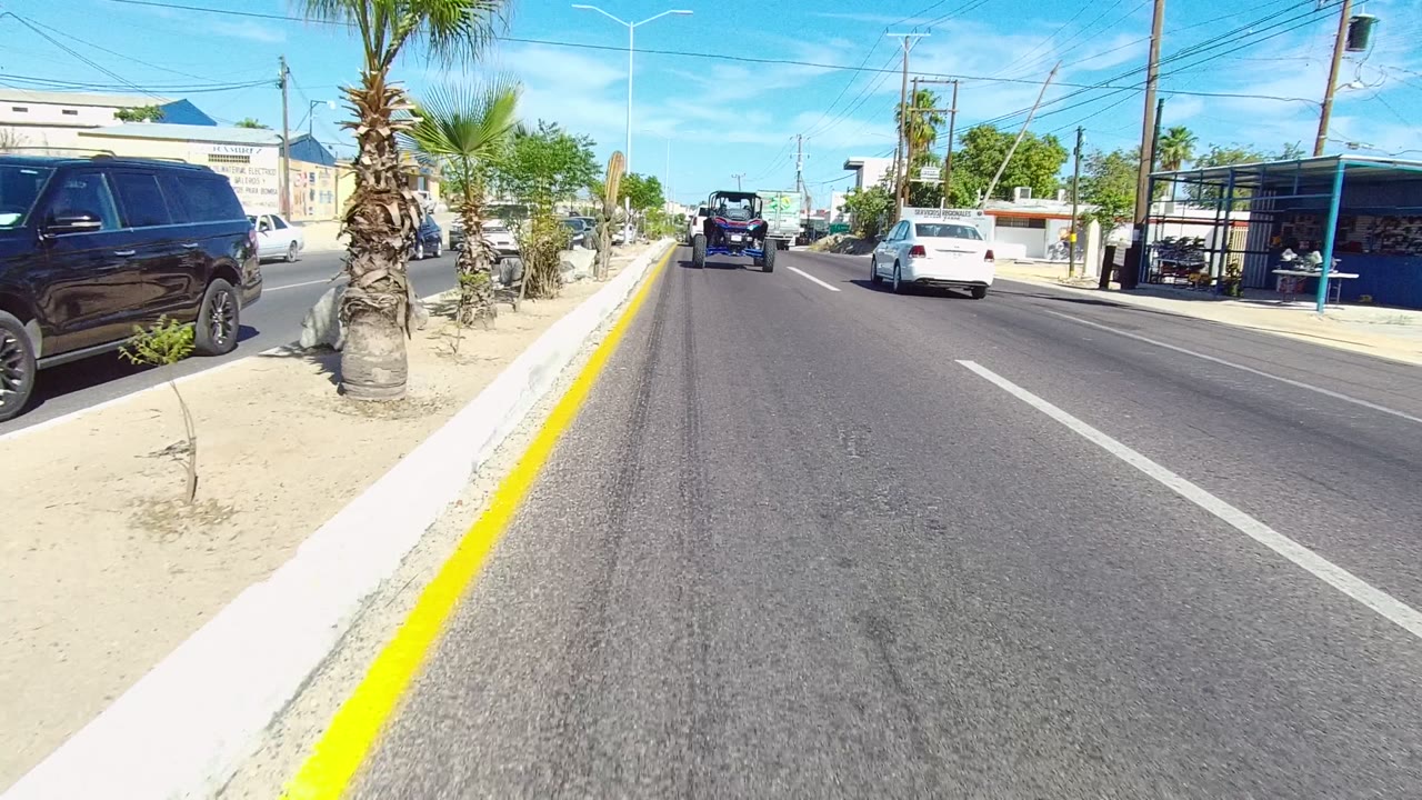 Start of a Los Cabos road view with a yellow curb line at left, parked vehicles on both sides, small palms, low buildings, and no visible people.