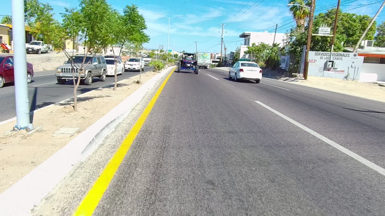 Mid-clip Los Cabos roadway view with cars ahead in the lane, a yellow curb line, roadside signs, sparse trees, low commercial buildings, and no visible people.
