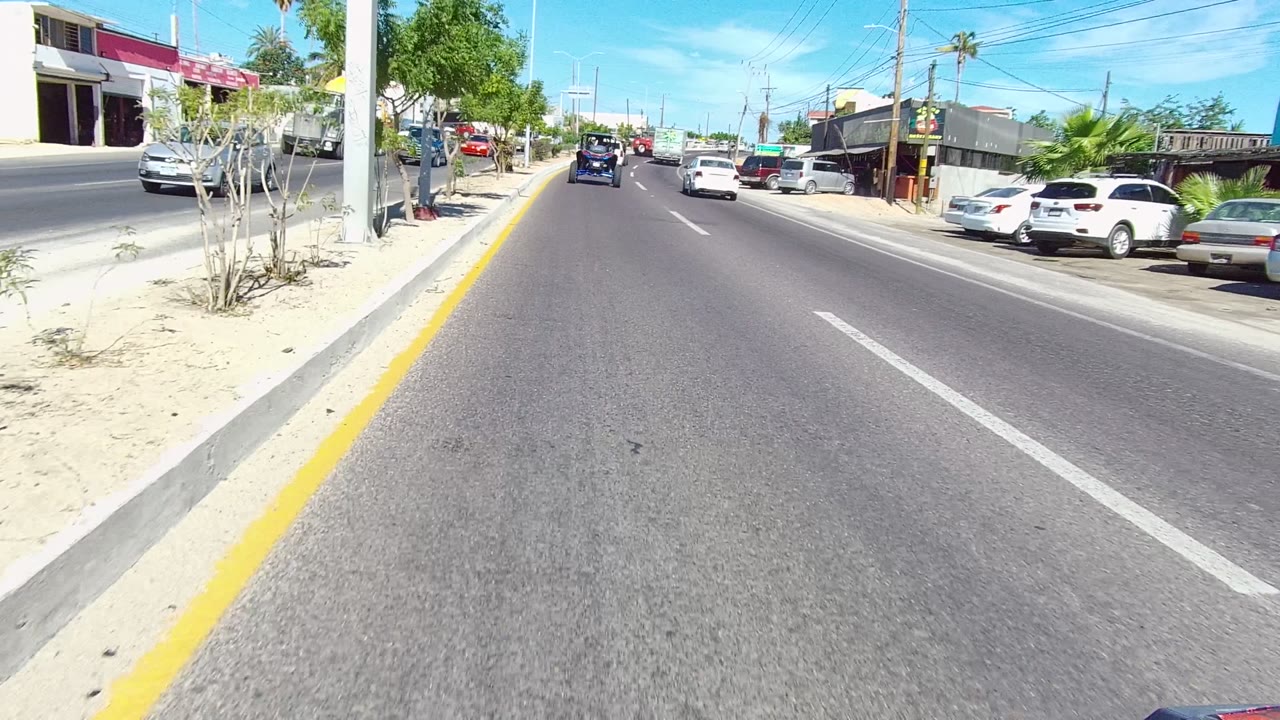 Sunny roadway with cars ahead and a row of parked vehicles beside buildings.