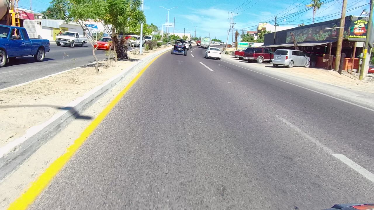Forward driving frame in Los Cabos showing the road, yellow median edge, a blue pickup truck raised on a roadside display or ramp, parked cars, businesses, trees, and no visible people.