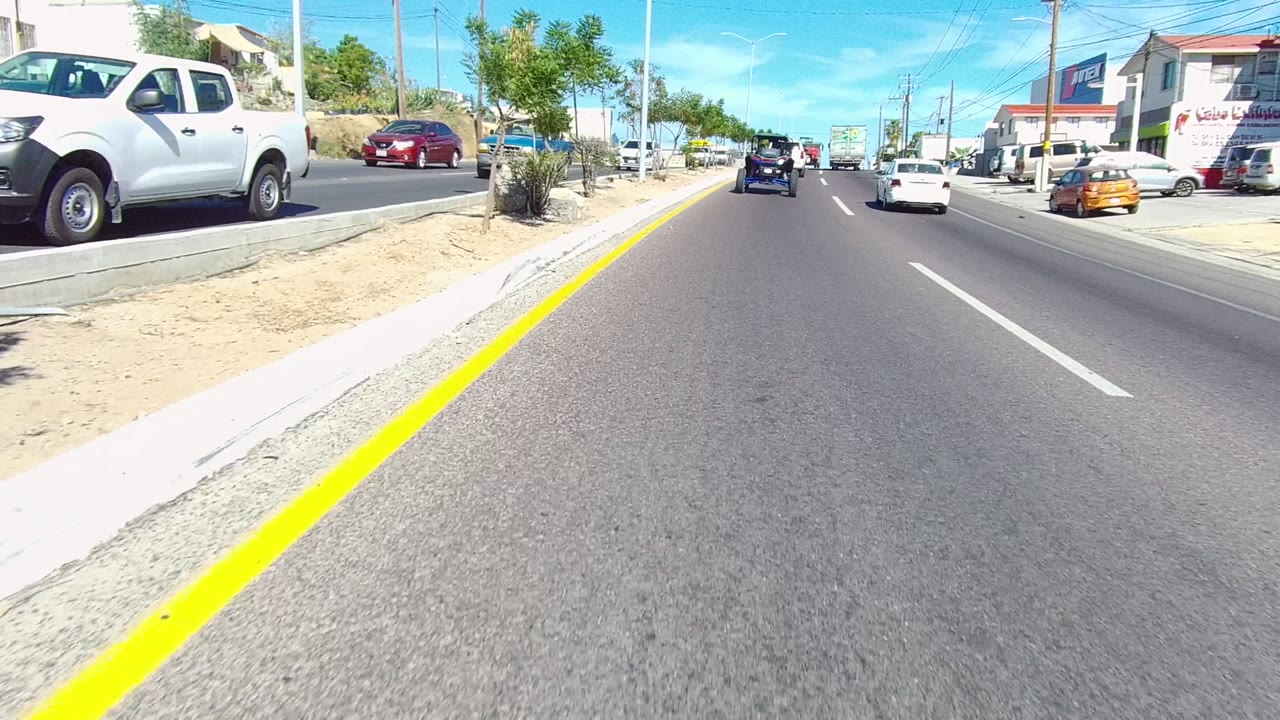 A forward road view in Los Cabos, Baja California Sur, Mexico shows cars and trucks on a sunny street with a yellow curb line, roadside trees, utility poles, commercial buildings, and no visible people.