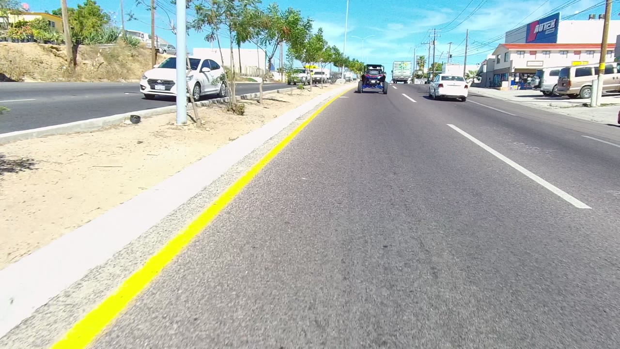 Road traffic passing roadside businesses with trees, parked vehicles, and clear sky.