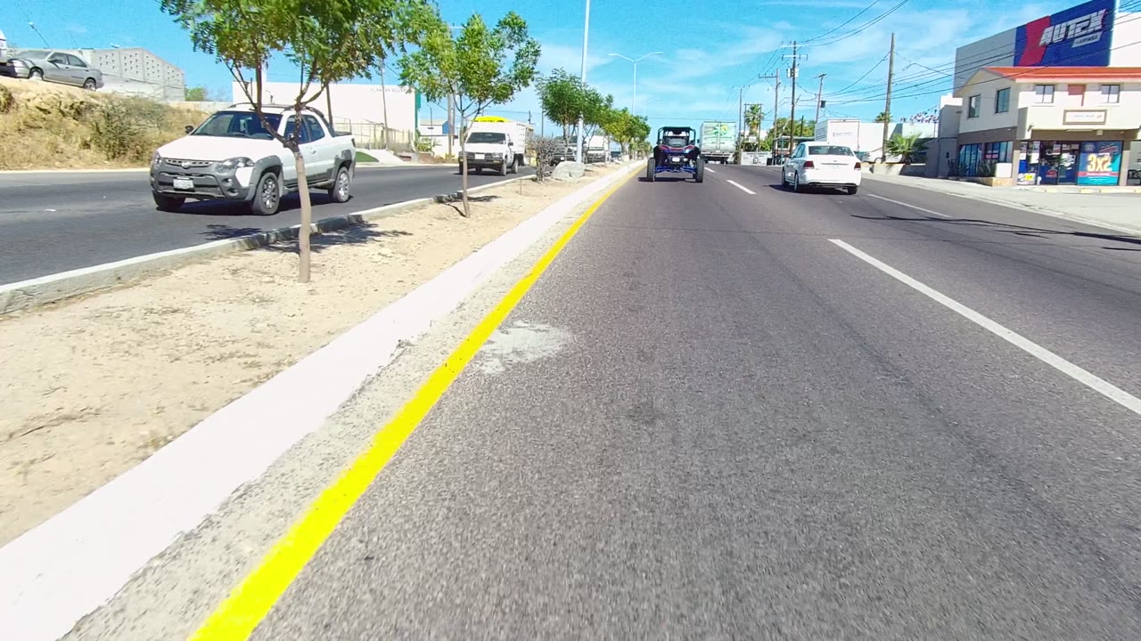 In Los Cabos, a pickup appears near the right side of the road while distant cars continue ahead, with curbside trees, commercial signage, utility poles, and no visible people.