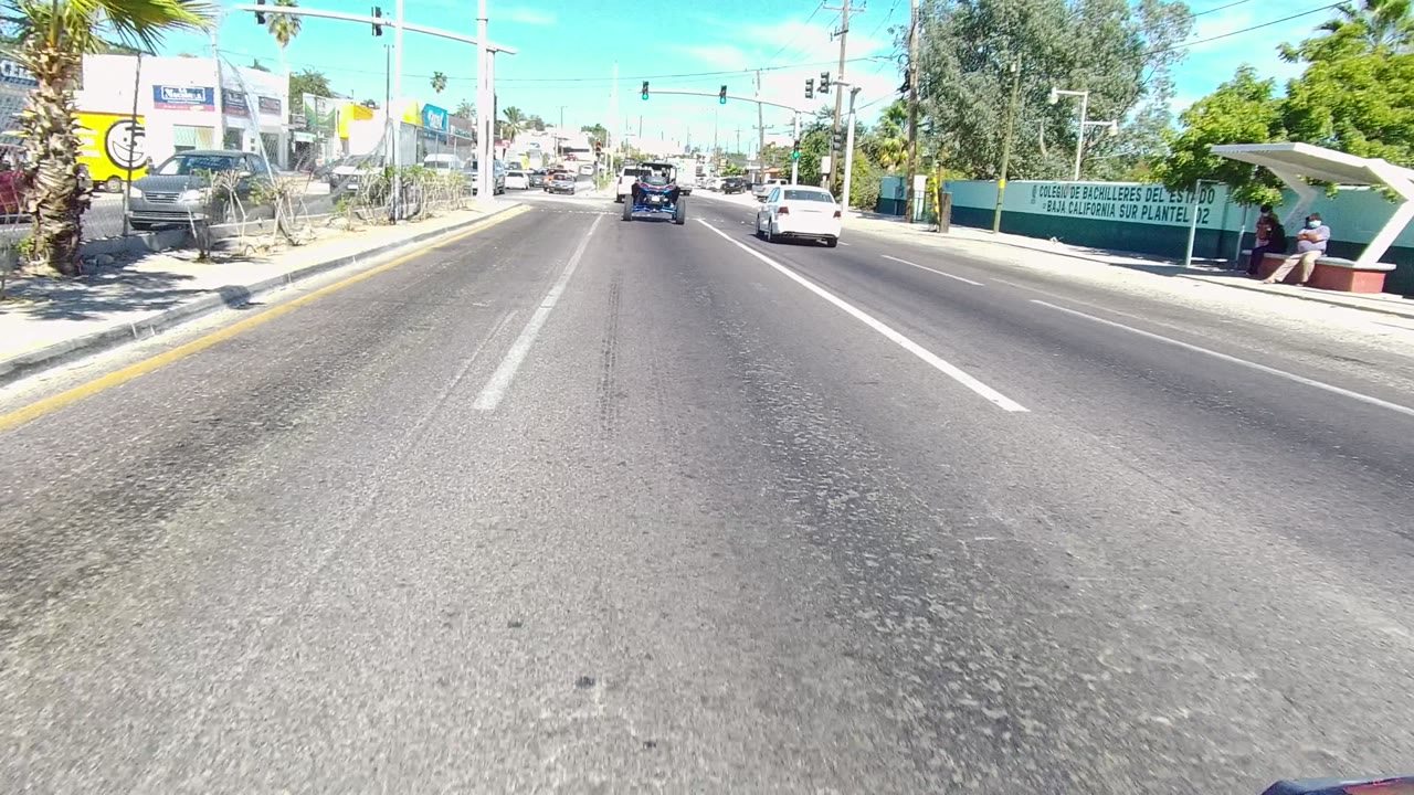 Forward road view in Los Cabos, Baja California Sur, showing cars ahead, traffic lights over the lanes, storefront signs, palm trees, and a roadside shelter with a possible distant person.