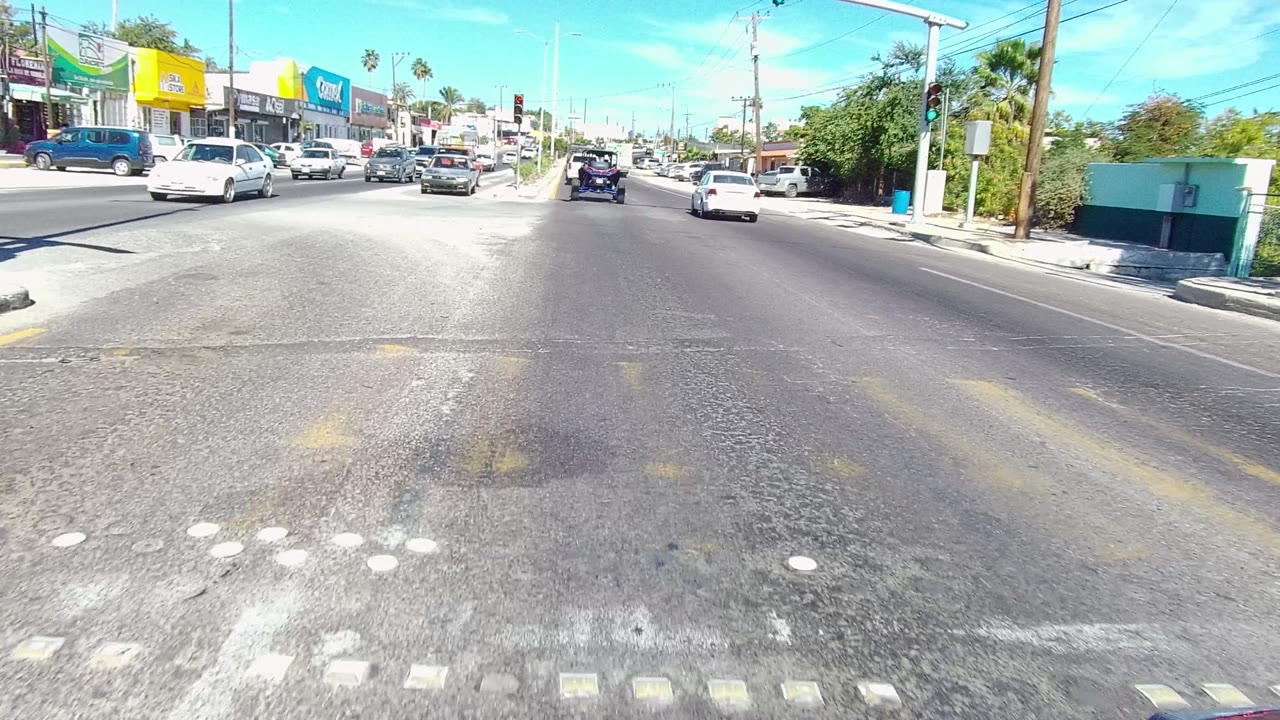 The road widens near an intersection in Los Cabos, with crosswalk markings in the foreground, cars in adjacent lanes, traffic signals, commercial signs, palm trees, and utility wires.
