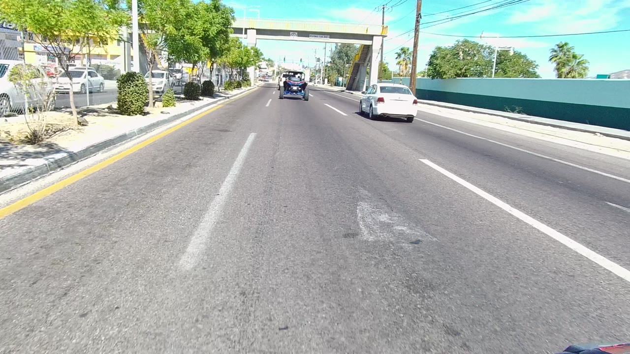 A forward road view in Los Cabos shows the overpass closer in frame, the white car moving along the right lane area, a blue vehicle farther ahead, and no visible people.