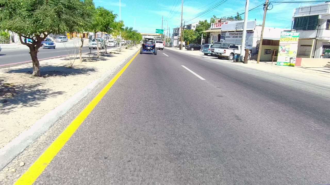 Start of the Los Cabos road clip, showing a paved street with a yellow center line, sandy sidewalk, trees, parked cars, low buildings, blue sky, and no visible people.